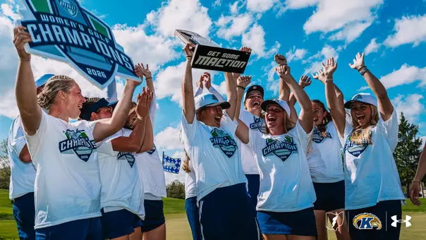 The Kent State women's golf team celebrates on the course, raising their arms and cheering while holding the 2026 MAC Women's Golf Champions trophy and a "Get Some MACtion" sign. The team wears white MAC Women's Golf Champions T-shirts and blue skirts. The Kent State and Under Armour logos appear in the bottom right corner.