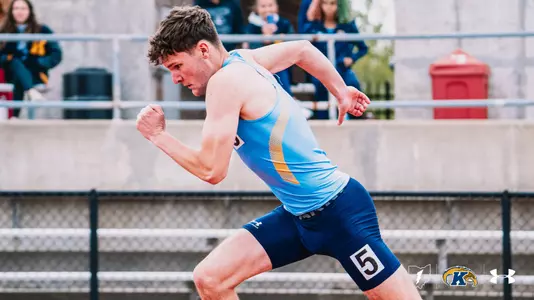 Kent State track and field athlete Preston Rieger, wearing a light blue Under Armour uniform with bib number 5 on his shorts, explodes out of the starting blocks during a sprint event at the OSU Invitational. The Kent State Athletics logo and Under Armour wordmark appear in the bottom right corner.