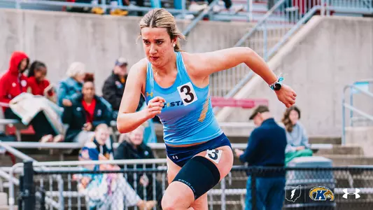 Kent State track and field athlete Ana Fernandez Moiron, wearing a light blue Under Armour uniform with "Kent State" across the chest and bib number 3, drives through a sprint race at the OSU Invitational, wearing a compression sleeve on her left leg. The Kent State Athletics logo and Under Armour wordmark appear in the bottom right corner.