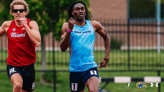 Kent State track and field athlete Micah Coates, wearing a light blue Under Armour uniform with "Kent State" across the chest and bib number 1, sprints ahead of a Miami (Ohio) competitor in red during a race at the OSU Invitational. The Kent State Athletics logo and Under Armour wordmark appear in the bottom right corner.