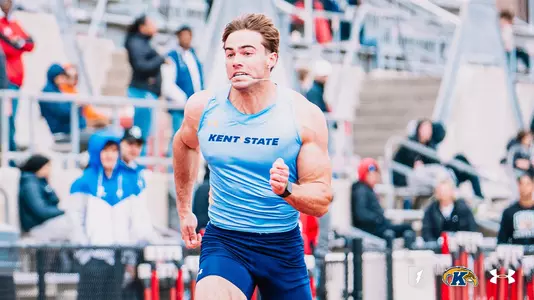 Kent State track and field athlete Ryan Gruss, wearing a light blue Under Armour uniform with "Kent State" across the chest, sprints during an event at the OSU Invitational, with hurdles visible along the track behind him. The Kent State Athletics logo and Under Armour wordmark appear in the bottom right corner.