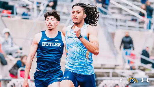 Kent State track and field Chaz Sakala, athlete wearing a light blue Under Armour uniform with "Kent State" across the chest, races ahead of a Butler competitor in a navy blue uniform during an event at the OSU Invitational. The Kent State Athletics logo and Under Armour wordmark appear in the bottom right corner.