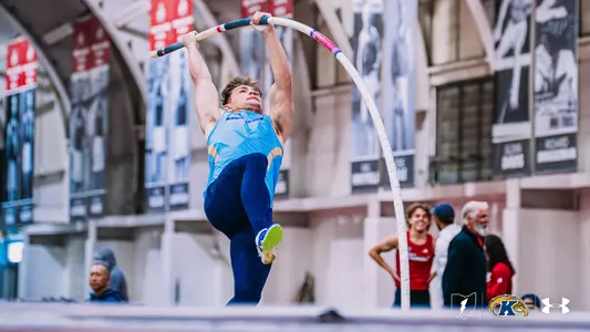 Kent State track and field athlete Braxton Quaintance, wearing a light blue Under Armour uniform with "Kent State" across the chest and navy tights, performs a pole vault attempt during an indoor event, gripping the pole at full extension as it bends beneath his weight. Championship banners and retired numbers are visible on the arena walls in the background. The Kent State Athletics logo and Under Armour wordmark appear in the bottom right corner.