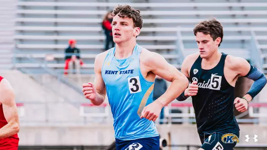 Kent State track and field athlete Owen Kelly, wearing a light blue Under Armour uniform with "Kent State" across the chest and bib number 3, runs during a distance event at the OSU Invitational. A competitor in a dark "Raiders" uniform with bib number 5 runs alongside him. The Kent State Athletics logo and Under Armour wordmark appear in the bottom right corner.