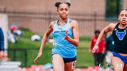Kent State track and field athlete Laila Eiland, wearing a light blue Under Armour uniform with "Kent State" across the chest and bib number 9, sprints during a race at the OSU Invitational. A competitor in a teal and blue Buffalo uniform runs to her right. The Kent State Athletics logo and Under Armour wordmark appear in the bottom right corner.