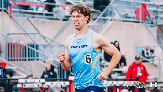 Kent State track and field athlete Caleb Bost, wearing a light blue Under Armour uniform with "Kent State" across the chest and bib number 6, runs during a distance event at the OSU Invitational, with hurdles visible in the background. The Kent State Athletics logo and Under Armour wordmark appear in the bottom right corner.