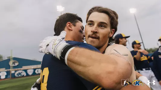 Two Kent State Golden Flashes baseball players embrace in celebration on the field, with teammates celebrating in the background, the Kent State Golden Flashes outfield wall visible behind them, and the Kent State lightning bolt, Golden Flashes, and Under Armour logos in the corner.