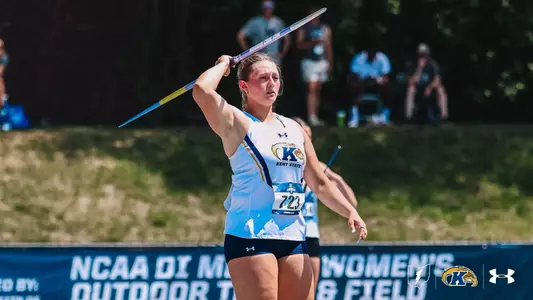Kent State javelin thrower Kristen Jay winds up for her throw at the NCAA Division I Men's and Women's Outdoor Track and Field Championships. Wearing bib No. 723 and a white Kent State Under Armour uniform, she holds the blue-and-yellow javelin above and behind her head, eyes focused forward. Spectators and a grassy embankment are visible in the background.