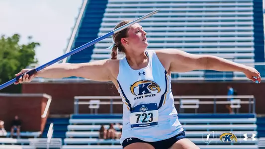 Kent State javelin thrower Kristen Jay extends into her release at the NCAA Championships in Lexington, Kentucky. Wearing bib No. 723 and a white Kent State Under Armour uniform, she stretches her arms wide — left hand gripping the blue javelin at full extension — as the implement climbs skyward. A stadium grandstand and blue seating structure fill the background.