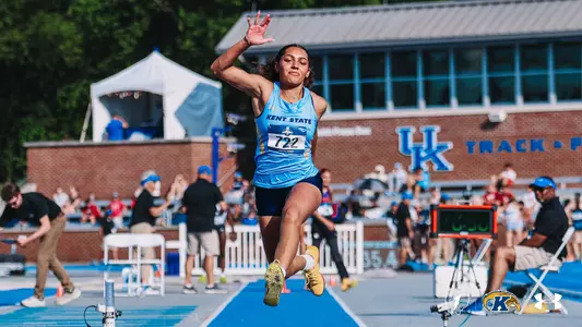 Kent State track and field athlete Lanee Hall, wearing bib No. 722, soars through the air during the long jump or triple jump at the University of Kentucky's track and field facility. She is dressed in a light blue Kent State singlet and navy shorts with gold spikes, her right arm raised and legs extended mid-flight. Spectators, officials, and timing equipment are visible in the background, with the UK Track & Field building prominently displayed behind her.