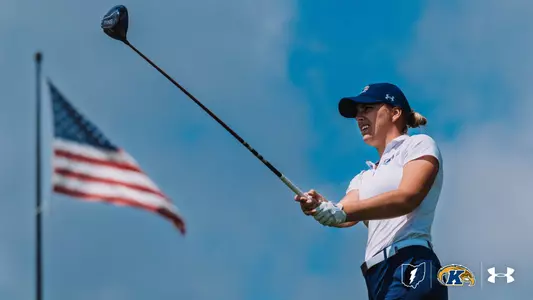 A Kent State women's golfer holds her follow-through and watches her drive against a bright blue sky, wearing a white Kent State polo, navy pants, a navy Under Armour cap, and a golf glove, with an American flag waving in the background and the Kent State lightning bolt, Golden Flashes, and Under Armour logos in the lower right corner.