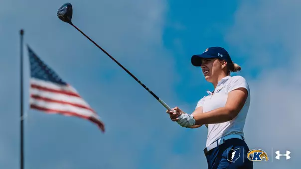 A Kent State women's golfer holds her follow-through and watches her drive against a bright blue sky, wearing a white Kent State polo, navy pants, a navy Under Armour cap, and a golf glove, with an American flag waving in the background and the Kent State lightning bolt, Golden Flashes, and Under Armour logos in the lower right corner.