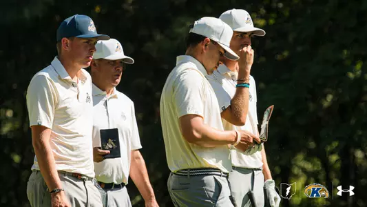Four Kent State men's golfers stand together in a line, studying the course ahead with focused expressions. All four wear matching cream and gold striped polo shirts and white caps with the Golden Flashes logo. Two players hold yardage books. A lush green tree line fills the background. The Kent State Golden Flashes logo and Under Armour wordmark appear in the lower right corner.