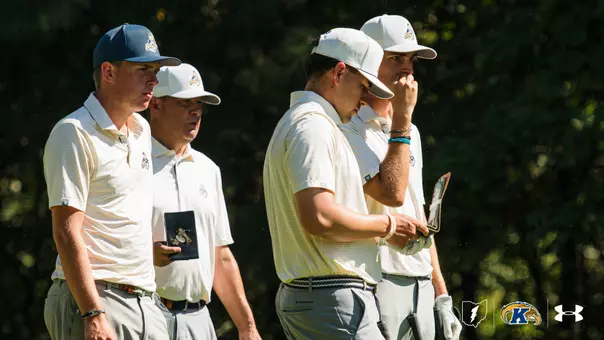Four Kent State men's golfers stand together in a line, studying the course ahead with focused expressions. All four wear matching cream and gold striped polo shirts and white caps with the Golden Flashes logo. Two players hold yardage books. A lush green tree line fills the background. The Kent State Golden Flashes logo and Under Armour wordmark appear in the lower right corner.
