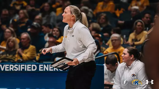 Kent State women's basketball head coach Fran Recchia stands on the sideline holding a clipboard, looking intently toward the court during a game.Kent State women's basketball head coach Fran Recchia stands on the sideline holding a clipboard, looking intently toward the court during a game. She wears a white Under Armour pullover with "Kent Basketball" lettering in gold and black pants, her blonde hair pulled back in a ponytail. A staff member in a matching white Kent State Under Armour jacket is seated behind her. The arena is filled with fans wearing gold and navy. A blue LED courtside advertising board reading "VERSIFIED ENER…" is visible in the background. The Kent State Golden Flashes logo, Ohio state outline, and Under Armour wordmark appear in the lower right corner.