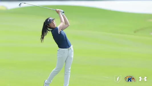 A Kent State women's golfer completes a full follow-through on an iron shot, wearing a navy polo, white pants, a white belt, and a navy Yonex visor, with her ponytail flowing behind her and a lush green fairway in the background, and the Kent State lightning bolt, Golden Flashes, and Under Armour logos in the lower right corner.