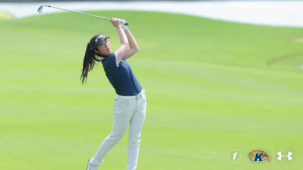 A Kent State women's golfer completes a full follow-through on an iron shot, wearing a navy polo, white pants, a white belt, and a navy Yonex visor, with her ponytail flowing behind her and a lush green fairway in the background, and the Kent State lightning bolt, Golden Flashes, and Under Armour logos in the lower right corner.
