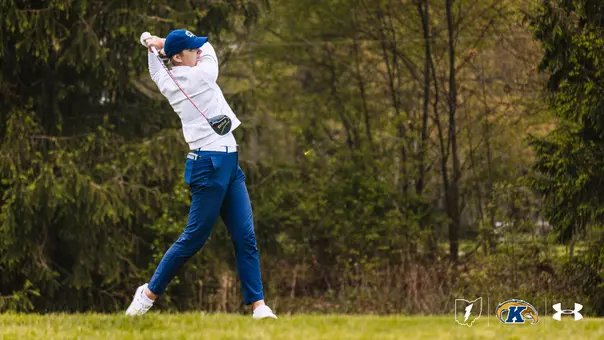A Kent State women's golfer completes a full follow-through on a driver swing, wearing a white long-sleeve top, blue pants, white shoes, and a blue Kent State cap, with a wooded course backdrop and the Kent State lightning bolt, Golden Flashes, and Under Armour logos in the lower right corner.