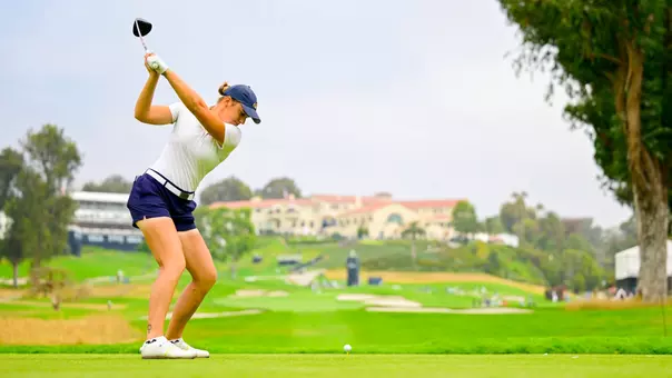 veronika kedVeronika Kedronova winds up at the top of her driver swing on the tee, wearing a white short-sleeve top, navy shorts, white Fila shoes, and a navy Kent State cap, with the ball on the tee in front of her, a lush tournament course, and a Spanish-style clubhouse visible in the background.onova at US Open