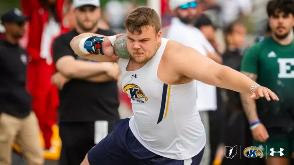 A Kent State track and field athlete competing in the shot put, wearing a white Under Armour singlet with the Kent State Golden Flashes logo and navy shorts. He grips the shot put against his neck with a taped right hand and braced wrist, his left arm extended back for balance during the throwing motion, with an intense, focused expression. Coaches, athletes, and spectators are visible blurred in the background, including an Eastern Michigan athlete in a green jersey. The Ohio state outline, Kent State Golden Flashes logo, and Under Armour logo appear in the bottom right corner.
