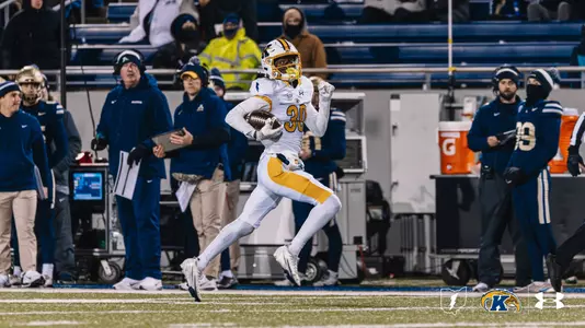 Kent State Golden Flashes football player wearing No. 30 in a white road uniform with gold accents runs along the sideline carrying the ball and pumping his fist, with the opposing team's coaches and players, a Gatorade cooler, and a stadium crowd visible in the background on a night under the lights.