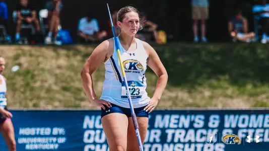 Kent State javelin thrower Kristen Jay stands with hands on hips, holding a javelin vertically at her side between throws, wearing a white Kent State Under Armour uniform and bib number 723. She gazes into the distance with a focused expression during the NCAA Men's and Women's Outdoor Track and Field Championships, hosted by the University of Kentucky. Spectators and a grassy hillside are visible in the background.