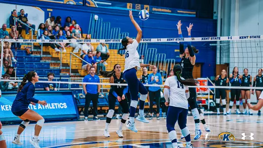 Kent State volleyball player Adalynn Ginley leaps to attack the ball at the net during a match against Buffalo, with a Buffalo blocker (No. 9) rising to defend as teammates from both squads look on from the court and bench.