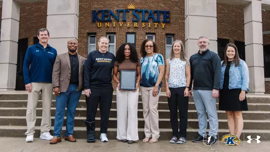 Dionna Gray holds an award plaque while standing with seven Kent State University administrators, coaches, and staff members on the steps in front of Kent State University's building entrance. A Kent State women's basketball coach stands to her left wearing a walking boot.