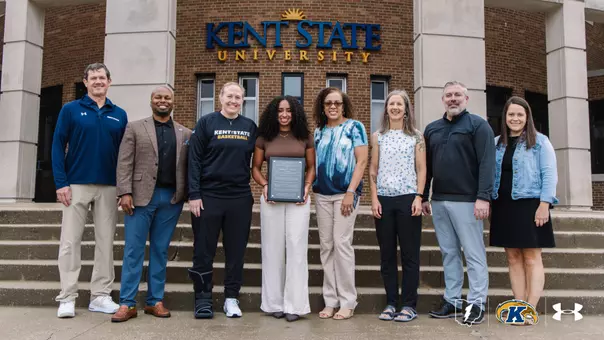 Dionna Gray holds an award plaque while standing with seven Kent State University administrators, coaches, and staff members on the steps in front of Kent State University's building entrance. A Kent State women's basketball coach stands to her left wearing a walking boot.