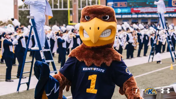 Flash, the Kent State Golden Flashes mascot, stands on the sideline in a navy 'Kent State' No. 1 football jersey, with the Kent State marching band performing in white and navy uniforms in the background and the Kent State lightning bolt, Golden Flashes, and Under Armour logos in the lower right corner.