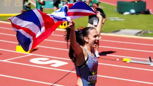 A track and field athlete wearing a Great Britain uniform celebrates while running with arms raised, waving a large Union Jack flag overhead. She has a dark ponytail with a red ribbon and is smiling with her mouth open in celebration. The scene is set on a red track with white lane markings, with a scoreboard and stadium officials visible in the blurred background.
