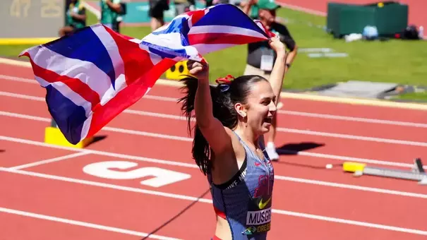A track and field athlete wearing a Great Britain uniform celebrates while running with arms raised, waving a large Union Jack flag overhead. She has a dark ponytail with a red ribbon and is smiling with her mouth open in celebration. The scene is set on a red track with white lane markings, with a scoreboard and stadium officials visible in the blurred background.