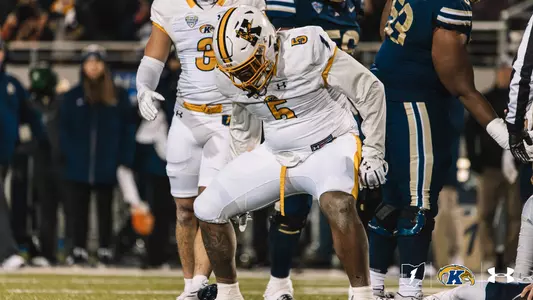 A Kent State football player wearing a white jersey with the number 5 and a white helmet stands on the field during a night game, looking down in a slightly crouched stance.