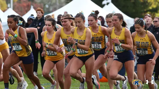 Kent State women's cross country runners sprint off the starting line at the beginning of a race, wearing gold Kent State singlets and navy shorts, with tents and spectators visible in the background.