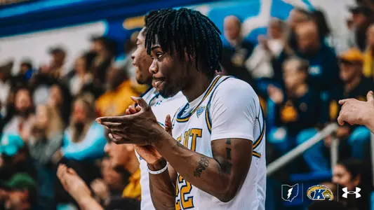 Kent State Men's Basketball player #22 Jaymen Townsend claps his hands in a white home jersey while on the court, with teammates and a crowd of fans cheering in the blurred background. The Kent State, and Under Armour logos appear in the bottom right corner.