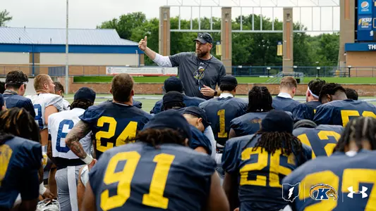 A football coach in a gray athletic shirt and cap gestures while addressing a gathered team of Kent State players on an outdoor practice field.