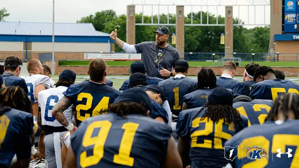 A football coach in a gray athletic shirt and cap gestures while addressing a gathered team of Kent State players on an outdoor practice field.