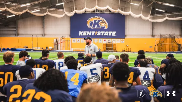 A football coach stands in an indoor athletic facility, addressing a group of Kent State football players seated on the turf in front of a large Kent State banner.