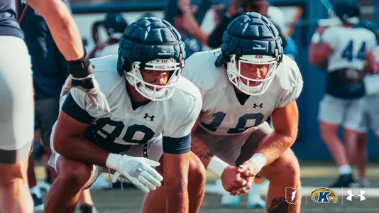 Two Kent State football players crouch side-by-side in a low stance during practice, wearing white practice jerseys and padded Guardian caps on their helmets.