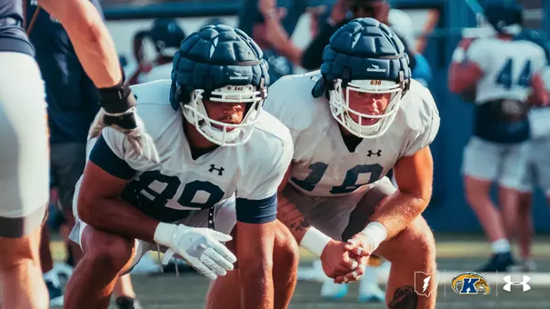 Two Kent State football players crouch side-by-side in a low stance during practice, wearing white practice jerseys and padded Guardian caps on their helmets.