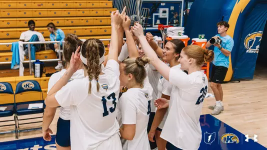 "Kent State volleyball players in white jerseys huddle together on the court, raising their joined hands overhead in celebration. In the background, gold bleachers, team staff, and Kent State branding line the arena."