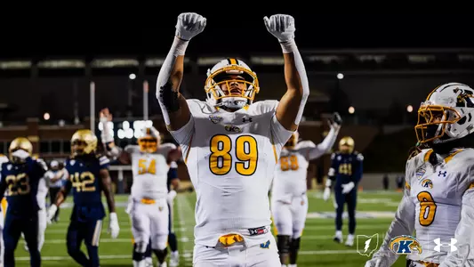 Kent State Golden Flashes player wearing No. 89 raises both fists in celebration on the field during a night road game, in a white road uniform with gold accents and a white helmet with gold facemask, with teammates and opposing players visible in the background and the Kent State lightning bolt, Golden Flashes, and Under Armour logos in the lower right corner.