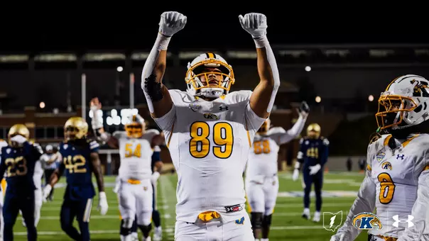 Kent State Golden Flashes player wearing No. 89 raises both fists in celebration on the field during a night road game, in a white road uniform with gold accents and a white helmet with gold facemask, with teammates and opposing players visible in the background and the Kent State lightning bolt, Golden Flashes, and Under Armour logos in the lower right corner.