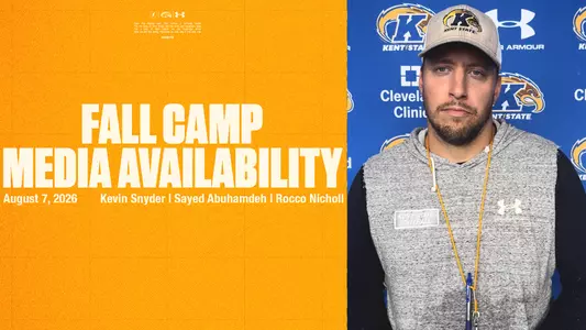 A split-screen graphic named FALL CAMP MEDIA AVAILABILITY 8.7.jpg. The left panel features a textured, bright yellow background with large, bold white text reading "FALL CAMP MEDIA AVAILABILITY." Below this, smaller white text says "August 7, 2026" and lists the names "Kevin Snyder | Sayed Abuhamdeh | Rocco Nicholl." Tiny logos and small print are visible at the very top of the yellow panel. The right panel displays a portrait of a man looking forward with a serious expression. He has short facial hair and is wearing a gray Kent State baseball cap, a gray sleeveless hoodie featuring a "Kent State Football" patch and an Under Armour logo over a black long-sleeve shirt, and a yellow lanyard. He stands against a blue step-and-repeat backdrop patterned with Kent State Athletics logos, as well as logos for the Cleveland Clinic and Under Armour.