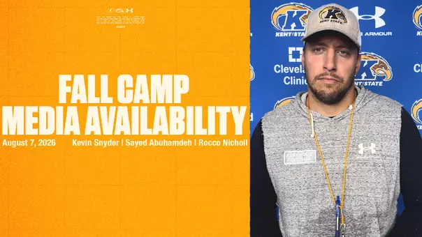A split-screen graphic named FALL CAMP MEDIA AVAILABILITY 8.7.jpg. The left panel features a textured, bright yellow background with large, bold white text reading "FALL CAMP MEDIA AVAILABILITY." Below this, smaller white text says "August 7, 2026" and lists the names "Kevin Snyder | Sayed Abuhamdeh | Rocco Nicholl." Tiny logos and small print are visible at the very top of the yellow panel. The right panel displays a portrait of a man looking forward with a serious expression. He has short facial hair and is wearing a gray Kent State baseball cap, a gray sleeveless hoodie featuring a "Kent State Football" patch and an Under Armour logo over a black long-sleeve shirt, and a yellow lanyard. He stands against a blue step-and-repeat backdrop patterned with Kent State Athletics logos, as well as logos for the Cleveland Clinic and Under Armour.