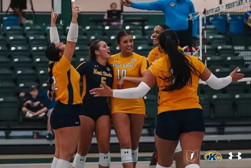 Five Kent State volleyball players celebrate on the court, wearing a mix of navy and gold jerseys, with arms raised and smiling. A referee in a blue shirt stands near the net in the background, with empty green stadium seating behind them.