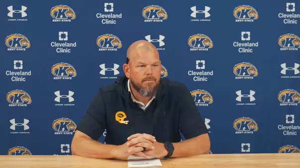 A man with a beard sits at a wooden table with hands clasped during a press conference in front of a blue Kent State backdrop.