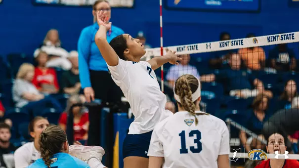 Kent State volleyball player No. 7 leaps to hit the ball at the net during a home match, with teammate No. 13 looking on and an opposing player attempting to block on the other side of the net.