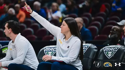 A Kent State Women's Basketball coach on the bench raises her right hand with a pen, signaling to players during a MAC Tournament game, as an assistant coach sits beside her.