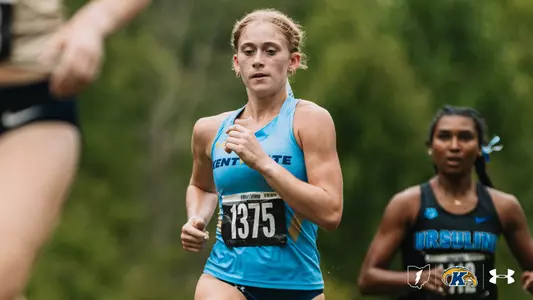 A Kent State cross country runner wearing bib No. 1375 competes in a race, running past a blurred competitor in the foreground. An Ursuline runner is visible behind her on the course.